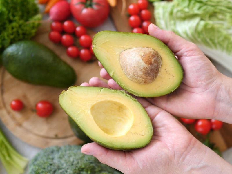 osoba otvara avokado, u pozadini daska sa paradajozom,. prokeljom i salatom