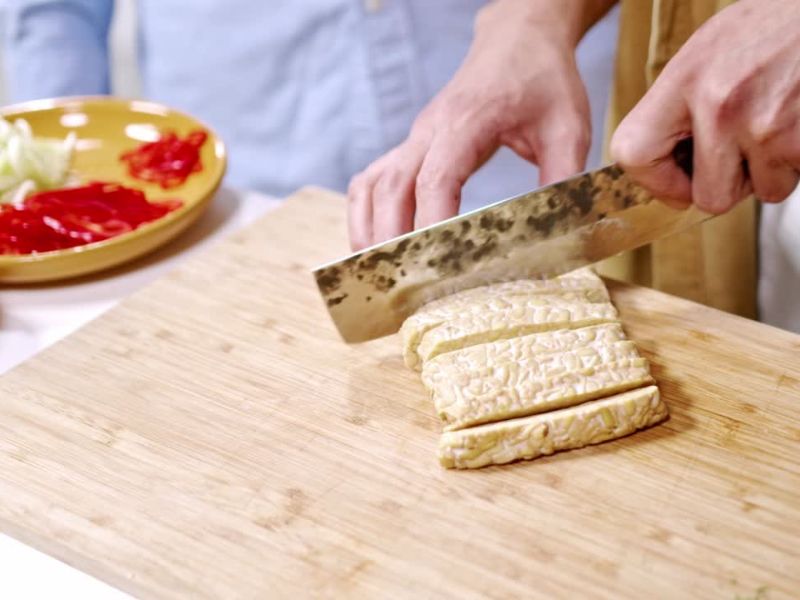 osoba seče tofu sir na dasci, pored nje tanjir sa povrćem